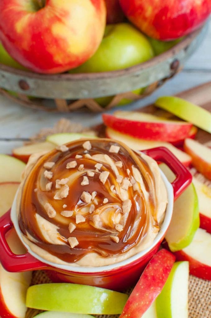 red bowl of caramel dip with caramel and toffee bits on top surrounded by sliced apples